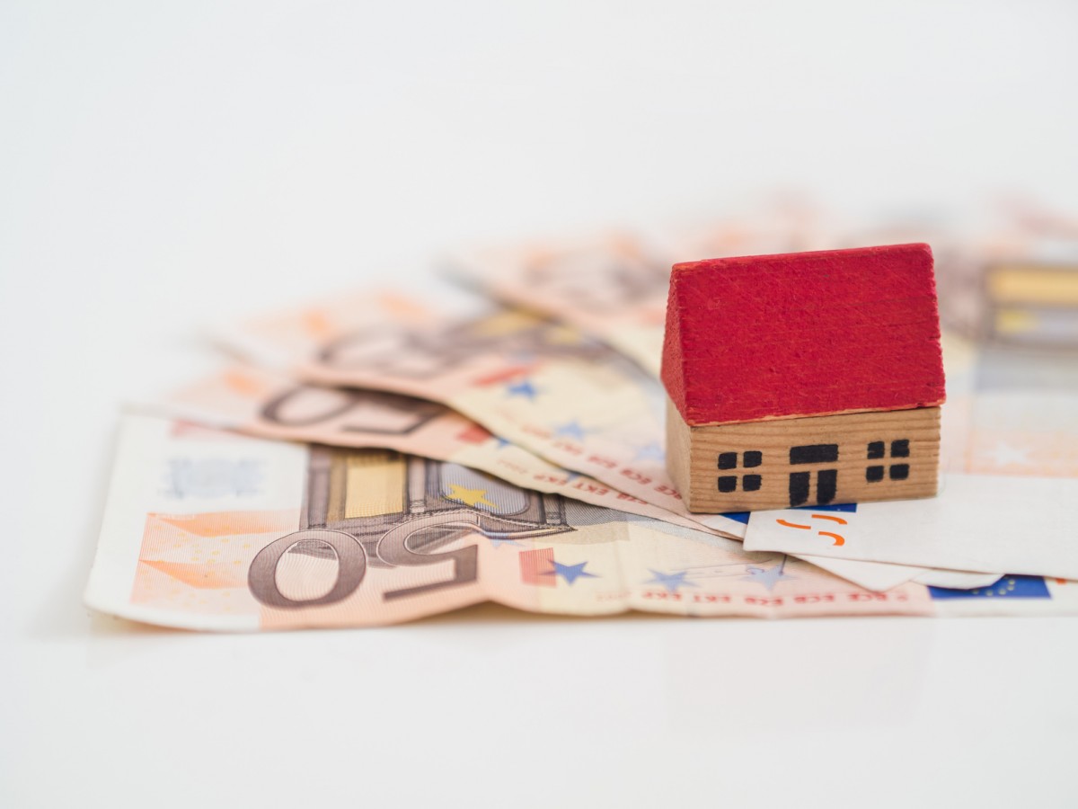 casita de juguete con tejado rojo sobre billetes de cincuenta euros en una mesa blanca.