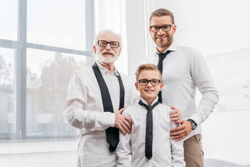 tres personas de diferente edad; abuelo, padre e hijo todos con corbata negra y camisa blanca. Al fondo, una ventana.