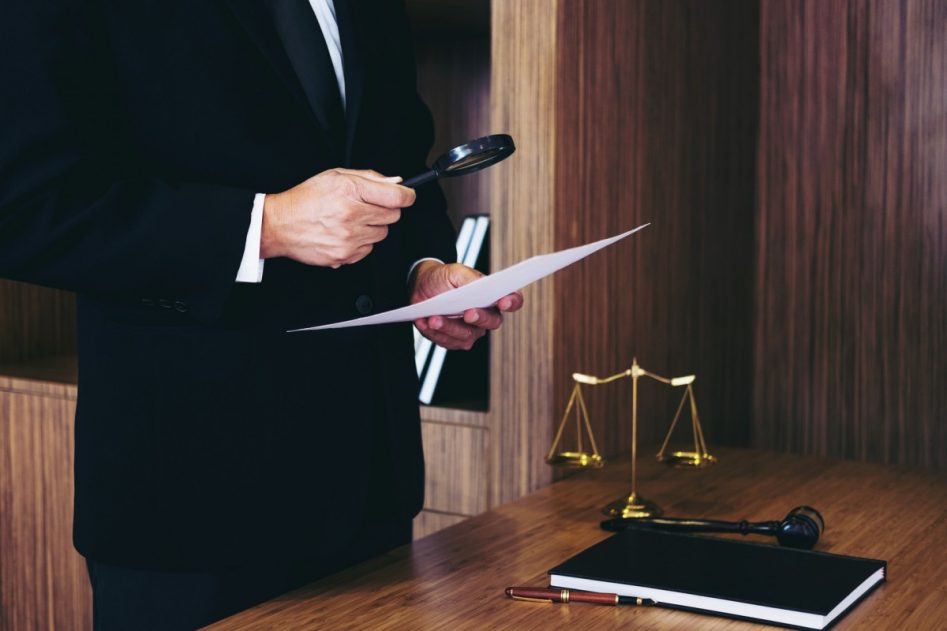 un hombre de pie, sostiene en sus manos y examina con una lupa un documento junto a una mesa de madera en la que hay una balanza legal, un martillo y un libro negro