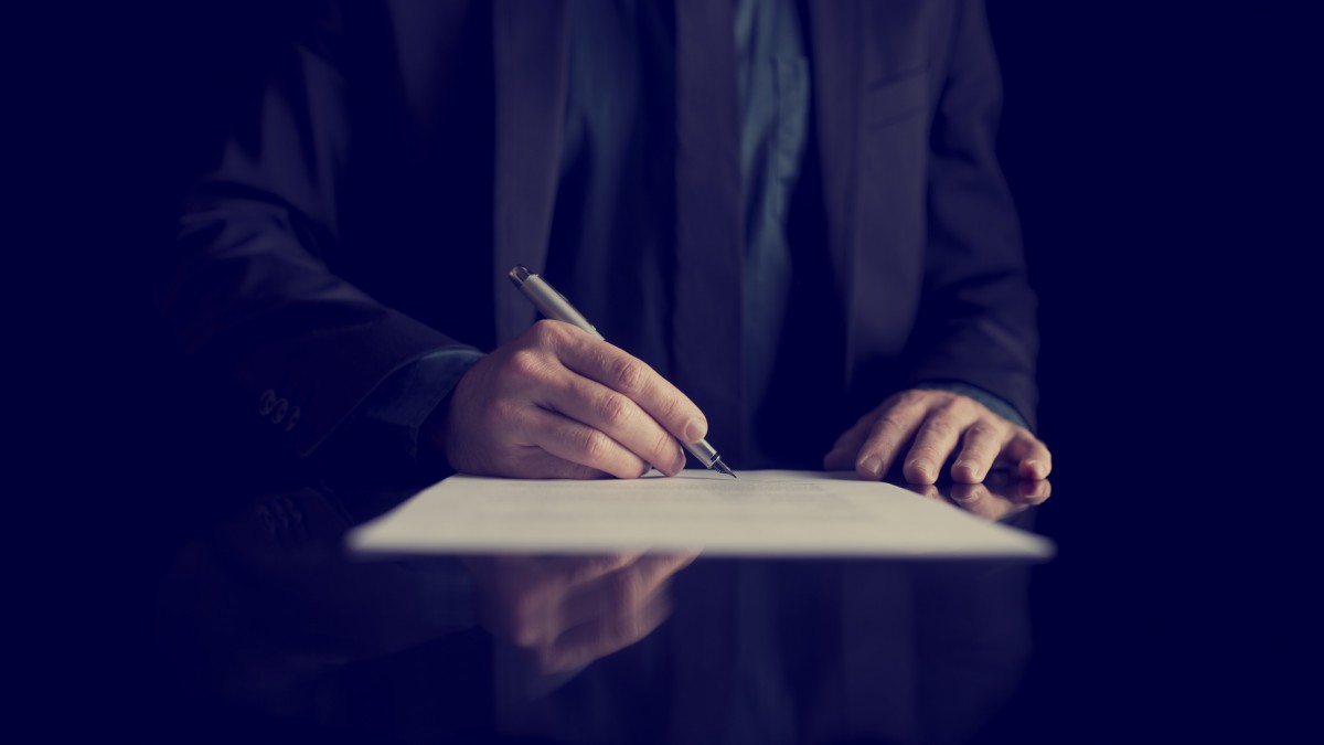 hombre con traje negro, firmando un documento sobre una mesa oscura